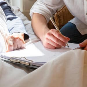 une main qui signe un document au chevet d'un mamalde. Comment bien remplir la clause bénéficaire de son assurance vie avec CPC & Associés